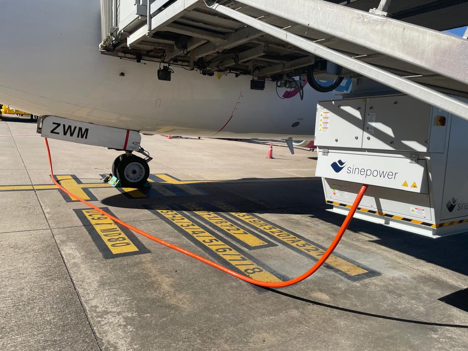 A Sinepower ground power unit on stand at an airport gate, cable connected to the aircraft